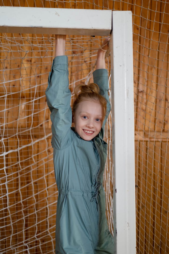 Young girl in green sportswear hanging on a net indoors, smiling at the camera.